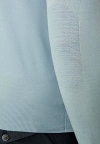 Světle modrý pletený svetr s rukávem, který má texturovanou nášivku na lokti a žebrovaný lem, přes tmavé kalhoty s viditelným knoflíkem.