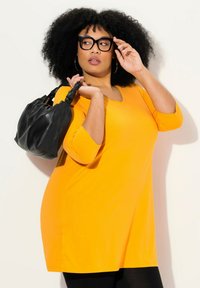 Übergröße orangefarbene Tunika mit drei Viertelärmeln, kombiniert mit schwarzen Leggings und einer lässigen schwarzen Handtasche. Glatter Stoff, legere Gestaltung.