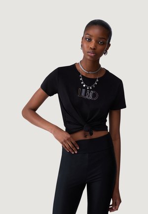 Giovane donna che indossa un top corto nero annodato con logo lucido, leggings neri, collane d'argento e orecchini a cerchio, posando con una mano sul fianco.