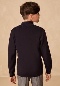 Tumesinise värvi kootud kampsun kõrge kraega ja pikkade varrukatega, koos hallide ruuduliste pükstega, tagantvaates neutraalse tausta ees.