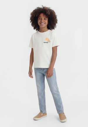 Niño sonriente con cabello rizado, vistiendo una camiseta blanca con estampado, jeans azul claro y zapatos beige sin cordones, de pie sobre un fondo blanco.