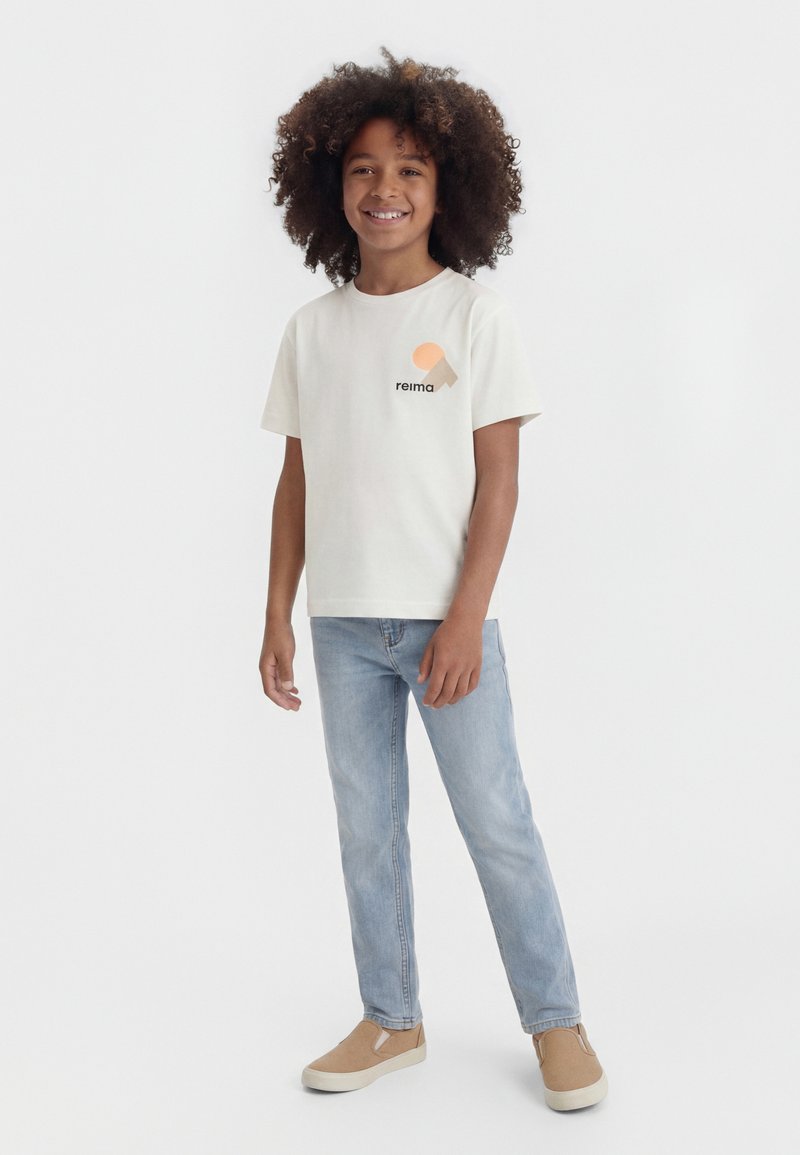 Χαμογελαστό παιδί με σγουρά μαλλιά, φορώντας λευκό μπλουζάκι με σχέδιο, ανοιχτό μπλε τζιν και μπεζ παπούτσια τύπου slip-on, στέκεται σε λευκό φόντο.