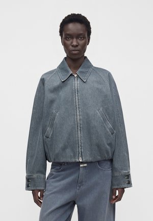 Personne portant une veste en jean bleu-gris oversize avec fermeture éclair et un pantalon ample assorti sur un fond clair uni.