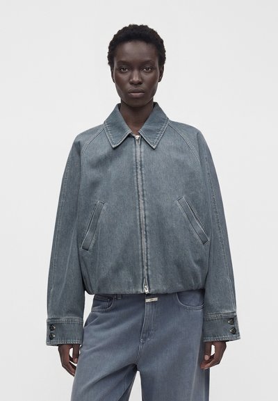 Personne portant une veste en jean bleu-gris oversize avec fermeture éclair et un pantalon ample assorti sur un fond clair uni.