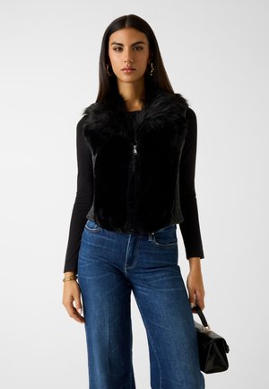 Zwart faux-fur gilet met ritssluiting, met een getextureerde achterkant, gecombineerd met blauwe wijde jeans en een kleine zwarte handtas.