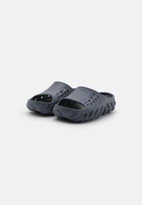 Mörkgrå slip-on klackar gjorda av mjukt material, med öppna sidor, ventilationshål och en texturerad sula med en vågformad design.