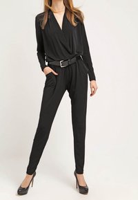 Combinaison noire à manches longues et décolleté en V, mettant en valeur une taille cintrée avec une ceinture et des jambes fuselées, assortie à des talons hauts noirs.