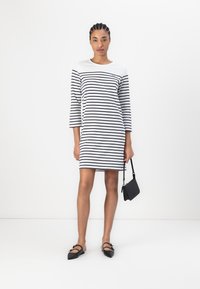 Rochie raiată în alb și bleumarin cu mâneci până la trei sferturi, decolteu rotund, formă ajustată și o geantă neagră tip crossbody. Pantofi negri cu talpă plată.