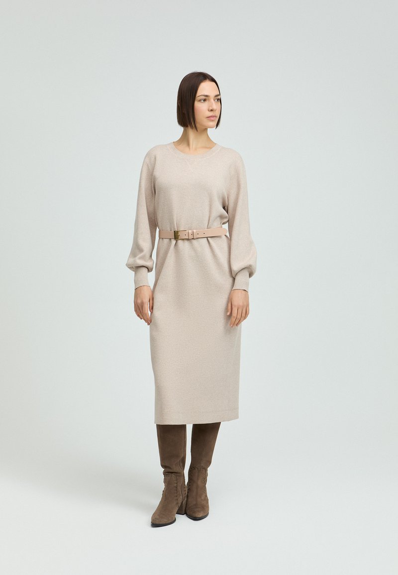 Beiges Strickkleid mit langen Puffärmeln und einer taillierten Taille, die von einem hellbraunen Gürtel betont wird. Kombiniert mit braunen kniehohen Stiefeln.