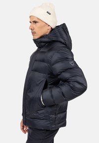 Giacca piumino di colore blu navy con colletto alto e chiusura con zip. Presenta un cappuccio e polsini elastici, con una toppa con logo sulla manica.