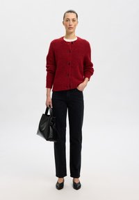 Sarkans adīts kardigāns ar melniem pogām, valkāts virs baltas krekla. Apvienots ar tumšām džinsām un melnu somu. Gluda tekstūra, ikdienišķs dizains.