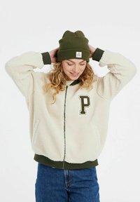 Krem žuti flis jakna sa zatvaranjem na zip i zelenim rebrastim manžetama, sa velikim izvezenim slovom "P" na grudima. Maslinasto zeleni beanie.