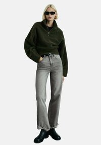 Olive zelen pulover iz flisa s pol-zadrgo, sproščenega kroja, v kombinaciji s sivimi hlačami visoke pasu in črnimi gležnjarji.