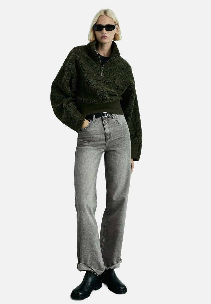 Olive zelen pulover iz flisa s pol-zadrgo, sproščenega kroja, v kombinaciji s sivimi hlačami visoke pasu in črnimi gležnjarji.