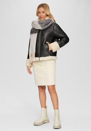 Zwarte leren jas met crèmekleurige faux shearling accenten, gecombineerd met een gestructureerde grijs-witte sjaal, crèmekleurige rok en beige enkellaarsjes.