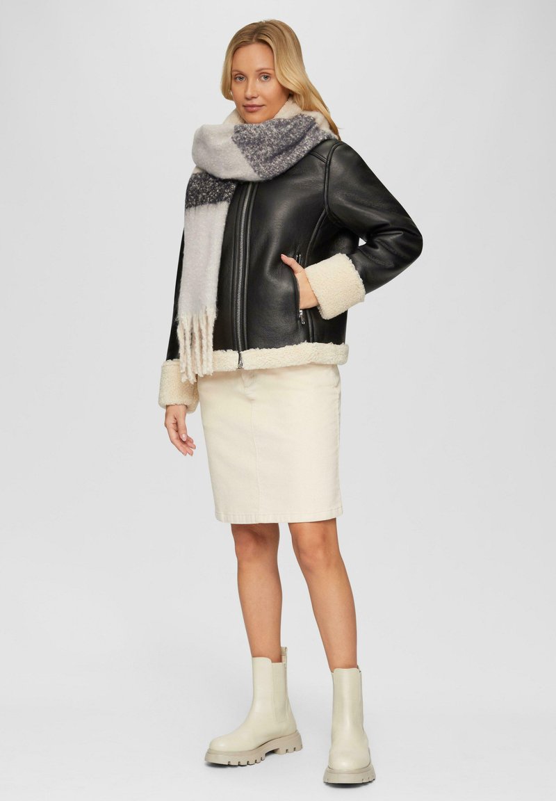 Veste en cuir noir avec des accents en fausse peau de mouton crème, associée à une écharpe texturée grise et blanche, une jupe crème et des bottines beige.
