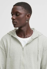 Felpa grigia chiara con zip, dal taglio rilassato, cappuccio con lacci e texture morbida; indossata sopra una t-shirt bianca, con una leggera lucentezza.