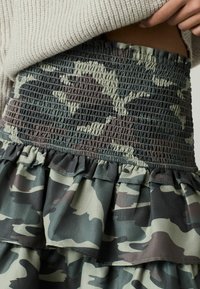 Jupe à volants avec une taille smockée en tissu camouflage dans des nuances de vert et de marron. L'ourlet froncé ajoute de la texture au design.