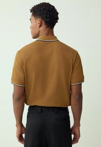 Camisa polo marrón con tela texturizada, cuello blanco a rayas, mangas cortas y combinada con pantalones negros. Vista desde la parte de atrás.