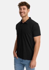 Polo negro de algodón con cuello clásico y mangas cortas, que presenta un pequeño logo tonal en el pecho. Combinado con unos vaqueros de color azul claro.