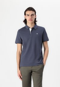Marinefarvet polo-shirts med korte ærmer, kontrasterende hvid kraveåbning og logo detaljer på brystet. Bæres med olivengrønne bukser.