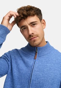 Pull bleu zippé avec col côtelé haut, tissu texturé et accent en cuir sur la tirette. Modèle regardant l'objectif, main dans les cheveux.