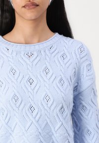 Hele sinine kudumipullover, millel on tekstuuriga teemanväär, avatud kirjutistega detailid, ümmargune kaelus ja sooniline mansett.