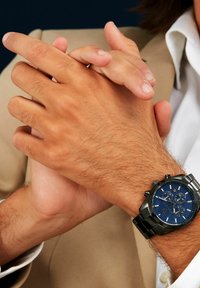 Herrklocka med svart metallarmband och blå urtavla, med tre subdials och datumsvisning. Händer sammanflätade mot en ljus tygbakgrund.