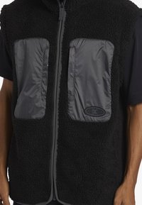 Svart fleeceväst med två framfickor i ett slätt grått material, dragkedjestängning fram och en logotyp på vänster ficka. Mjuk textur.