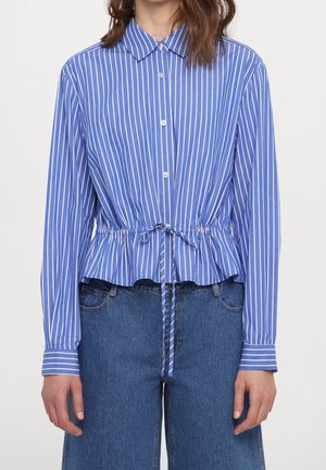 Kobieta ubrana w niebiesko-białą pionowo prążkowaną koszulę na guziki z troczkiem w talii i długimi rękawami, połączoną z niebieskimi dżinsami.