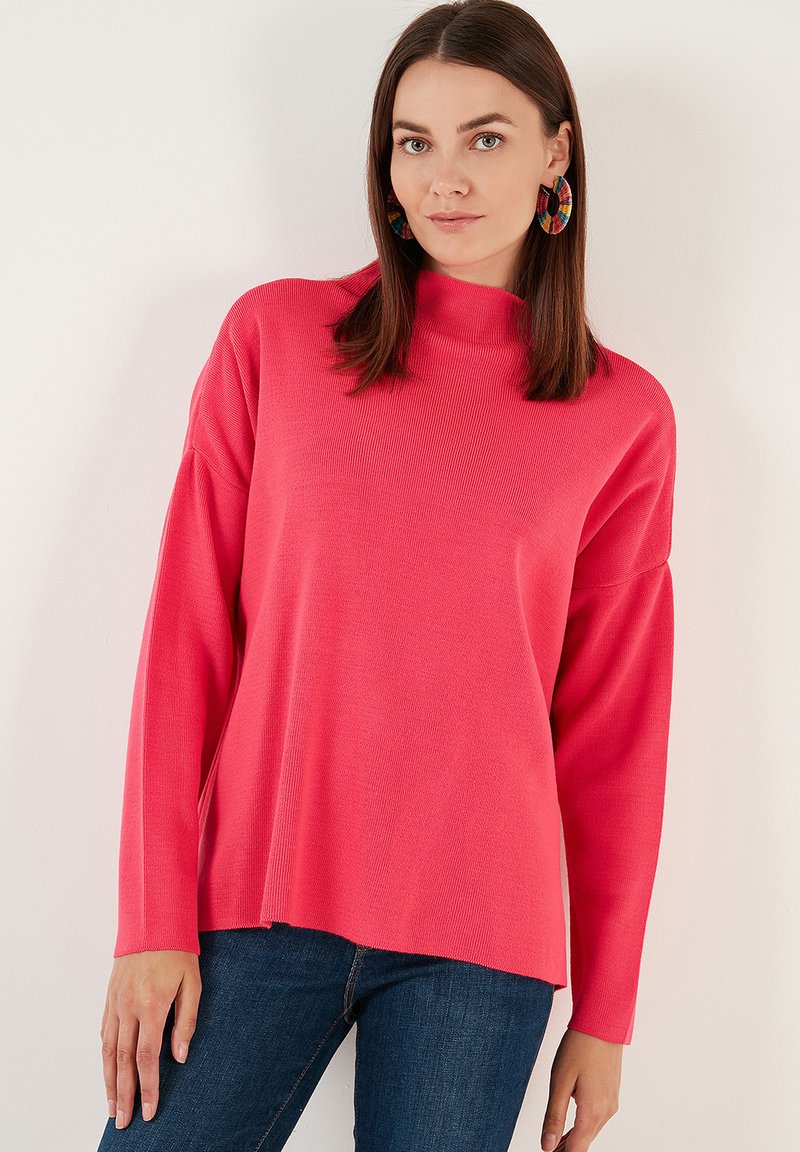 Pull rose à manches longues avec col montant, texture côtelée et coupe décontractée, associé à un jean en denim noir. La couleur est vive et uniforme.