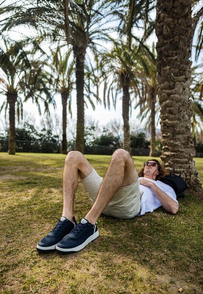 Des baskets bleu marine avec des semelles blanches présentent un dessus en tissu texturé et un système de laçage. Portées avec un short beige clair sur de l'herbe sous des palmiers.