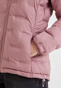 Light pink puffer jacket with a quilted design, elastic cuffs, and side pockets. Adjustable hem detail with drawstring.