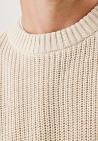 Pull en tricot couleur crème avec un col côtelé, présentant un motif texturé et une épaisseur moyenne.