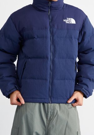 Mann trägt eine dunkelblaue The North Face Steppjacke mit Frontreißverschluss und graue Cargohosen, Hände richten den Saum der Jacke.