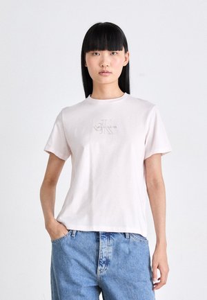 Jongere met steil zwart haar, gekleed in een lichtroze Calvin Klein T-shirt en blauwe jeans, staand tegen een effen witte achtergrond.