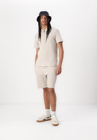 Beige strikket polo med korte ærmer og matchende shorts, som har en elastisk talje, parret med hvide sneakers og en mørk bucket hat.