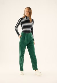 Green tailored trousers with pleats, side pockets, and button accents. Paired with a black and white striped turtleneck. Casual white sneakers.