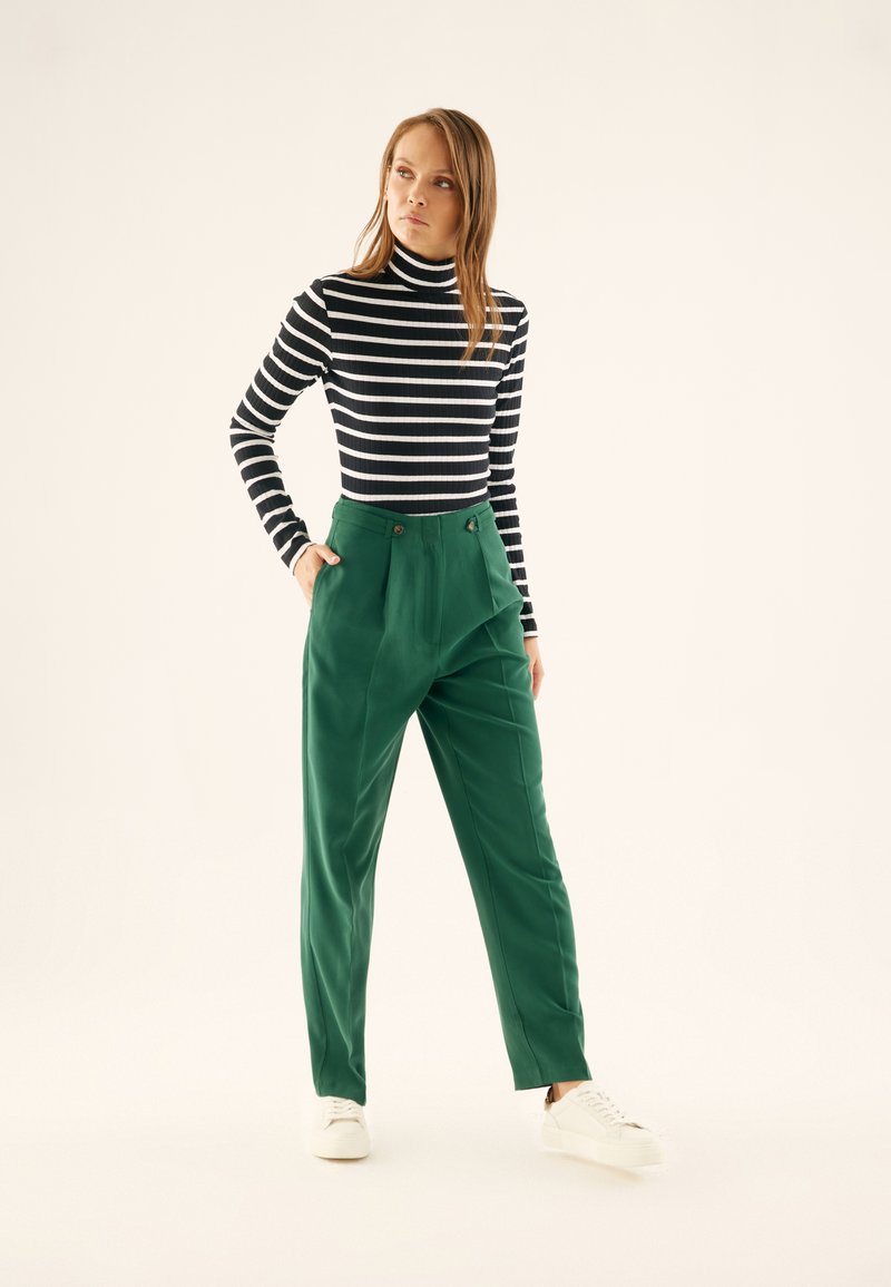 Green tailored trousers with pleats, side pockets, and button accents. Paired with a black and white striped turtleneck. Casual white sneakers.