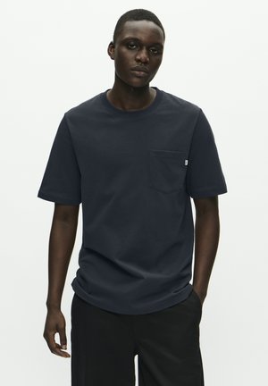 Ung man med mörk hud som bär en marinblå pocket-t-shirt och svarta byxor, står mot en helt ljus bakgrund.
