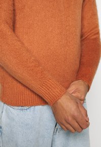 Pomarańczowy dziergany sweter z długimi rękawami i prążkowanymi mankietami, o miękkiej fakturze. W połączeniu z jasnoniebieskimi jeansami.