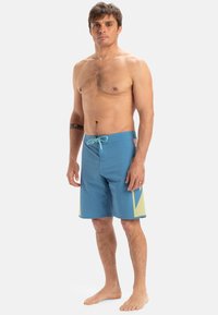 Modré plavky s hladkou, ľahkou textúrou. Majú kontrastný žltý geometrický vzor na stranách a šnúrkový uzáver.