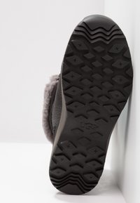 Suela de goma negra con un diseño texturizado y agarres, que muestra un upper de piel de oveja gris y un suave forro interior. Logotipo de UGG visible.