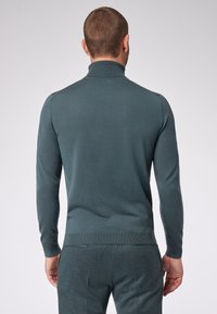 Von hinten sichtbarer Mann in einem eng anliegenden türkisfarbenen Rollkragenpullover und einer passenden Hose vor einem schlichten, hellen Hintergrund.