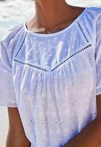 Weiße Baumwollbluse mit Stickdetails, die florale Muster, Augenlash-Ausschnitte und eine lässige Passform aufweist, ergänzt durch einen runden Ausschnitt.