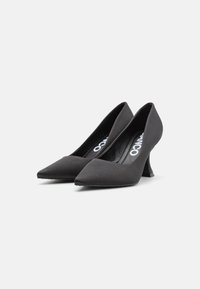 Zapatos de tacón negro de punta afilada con una textura similar al ante, que presentan un tacón angular distintivo y un diseño suave y minimalista.