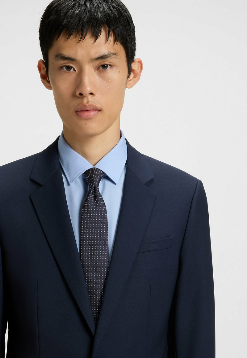 Giacca da abito blu navy con rever a punte, abbinata a una camicia azzurra chiara e una cravatta scura a motivo. Tessuto liscio, design ben aderente.