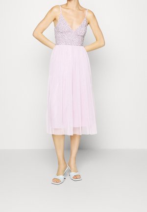 Vrouw draagt een lichtroze midi-jurk met een met kraaltjes versierde lijf en dunne bandjes, gecombineerd met witte open-teen sandaal met hak.