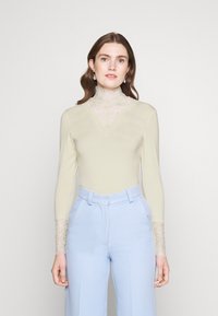 Krämfärgad långärmad bodysuit med spetskrage och spetsdetaljer vid ärmsluten, tillsammans med ljusblå höga byxor.