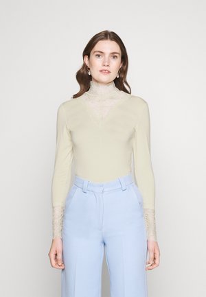 Cremfarbener Langarm-Body mit einem Spitzenrollkragen und Spitzenakzenten an den Manschetten, kombiniert mit hellblauen hochtaillierten Hosen.